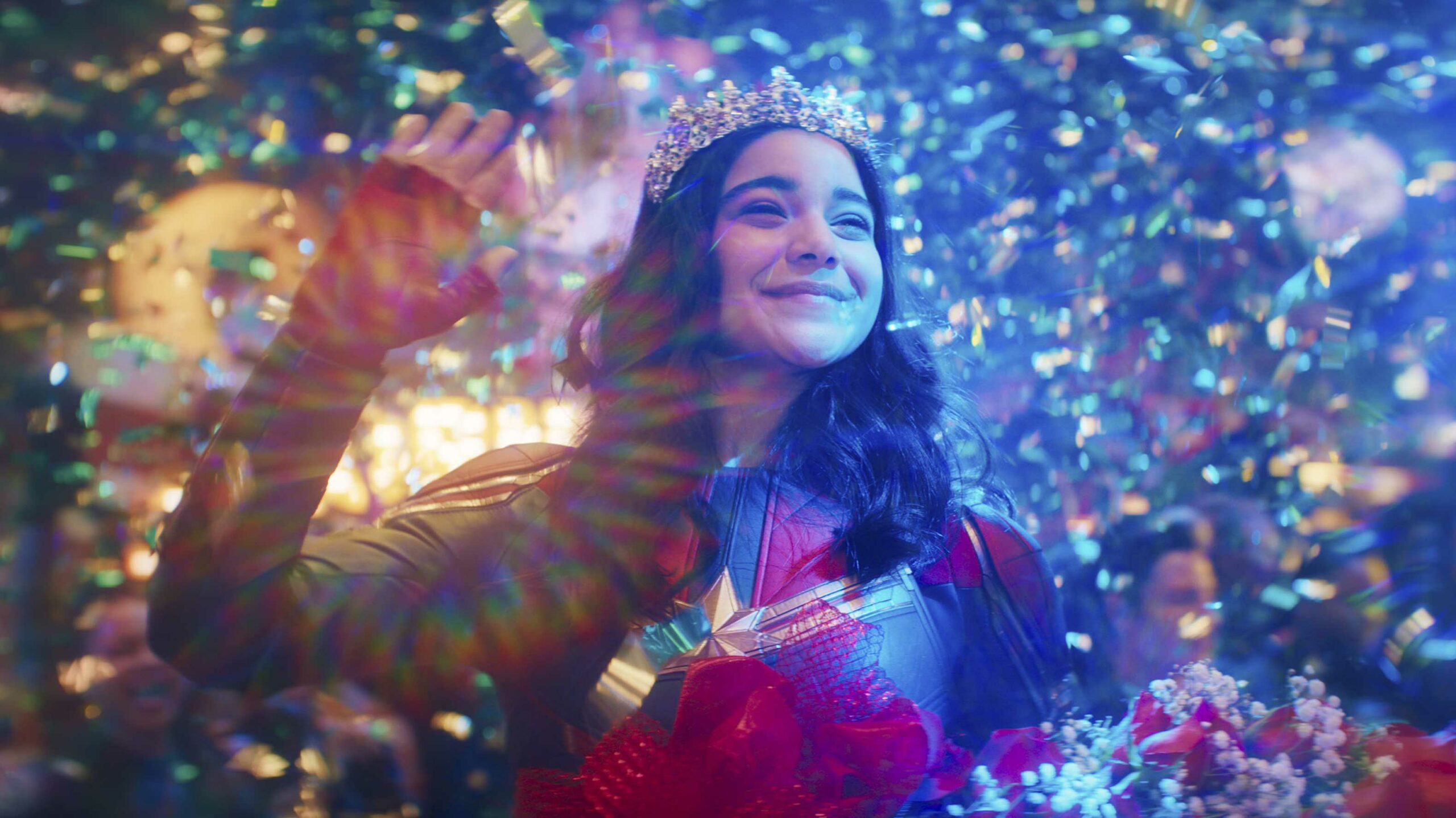 Iman Vellani as Kamala Khan/Ms. Marvel in the Disney+ series of the same name. She's smiling and waving as she receives flowers and confetti rains around her.